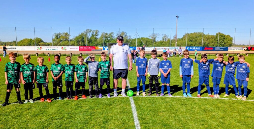 Aufstellung beider Mannschaften vor dem Anpfiff beim Auswärtsspiel der DJK Straubing U9 in Bogen – die Kinder in grün-schwarzen und blauen Trikots posieren gemeinsam mit dem Schiedsrichter auf dem Hauptspielfeld, im Hintergrund die Werbebanden der Sponsoren.