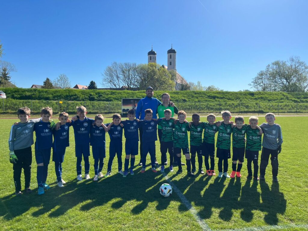 Mannschaftsfoto der DJK Straubing U9 gemeinsam mit dem Trainerteam und dem SC Kirchroth nach dem Spiel auf dem Sportplatz in Kirchroth, im Hintergrund die markante Pfarrkirche St. Margaretha bei strahlendem Frühlingswetter.