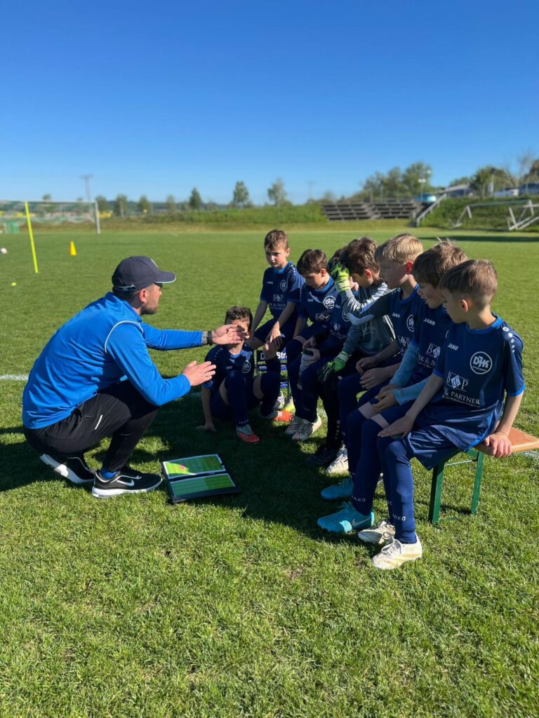 Trainer der DJK Straubing erklärt seinen U9-Spielern bei der Halbzeitpause taktische Anweisungen mithilfe einer Taktiktafel – konzentrierte Atmosphäre auf dem Trainingsplatz bei Sonnenschein.